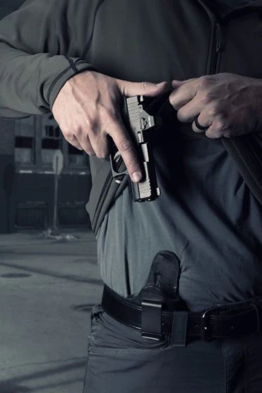 Close-up of a man’s waist showing a black IWB holster seated deep inside the pants with the clip attached over a black belt, while he lifts his shirt and draws a handgun.